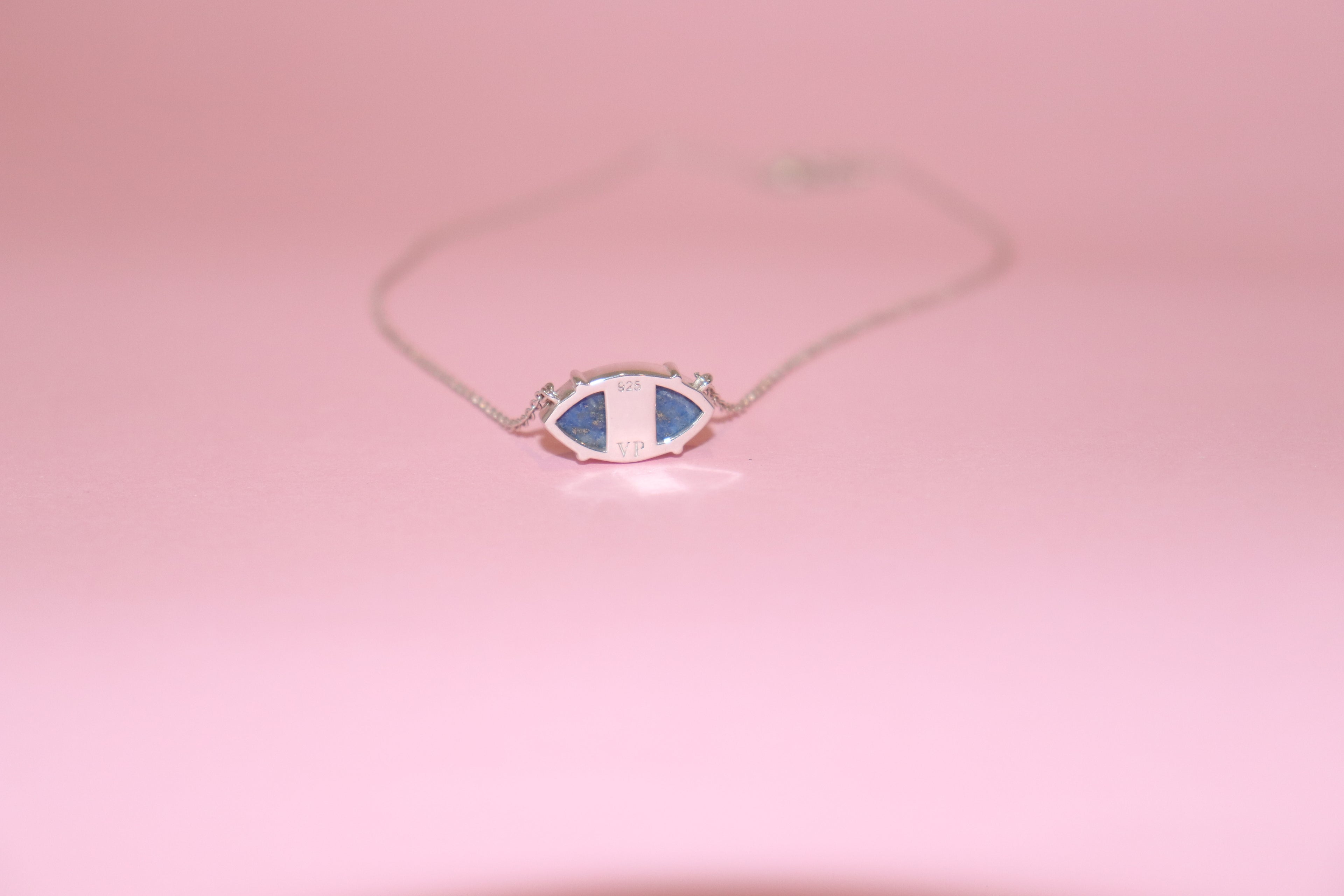 Silver necklace sitting on pink surface with a marquise pendant engraved with "925 VP", and set with blue lapis lazuli stone and on a pink background
