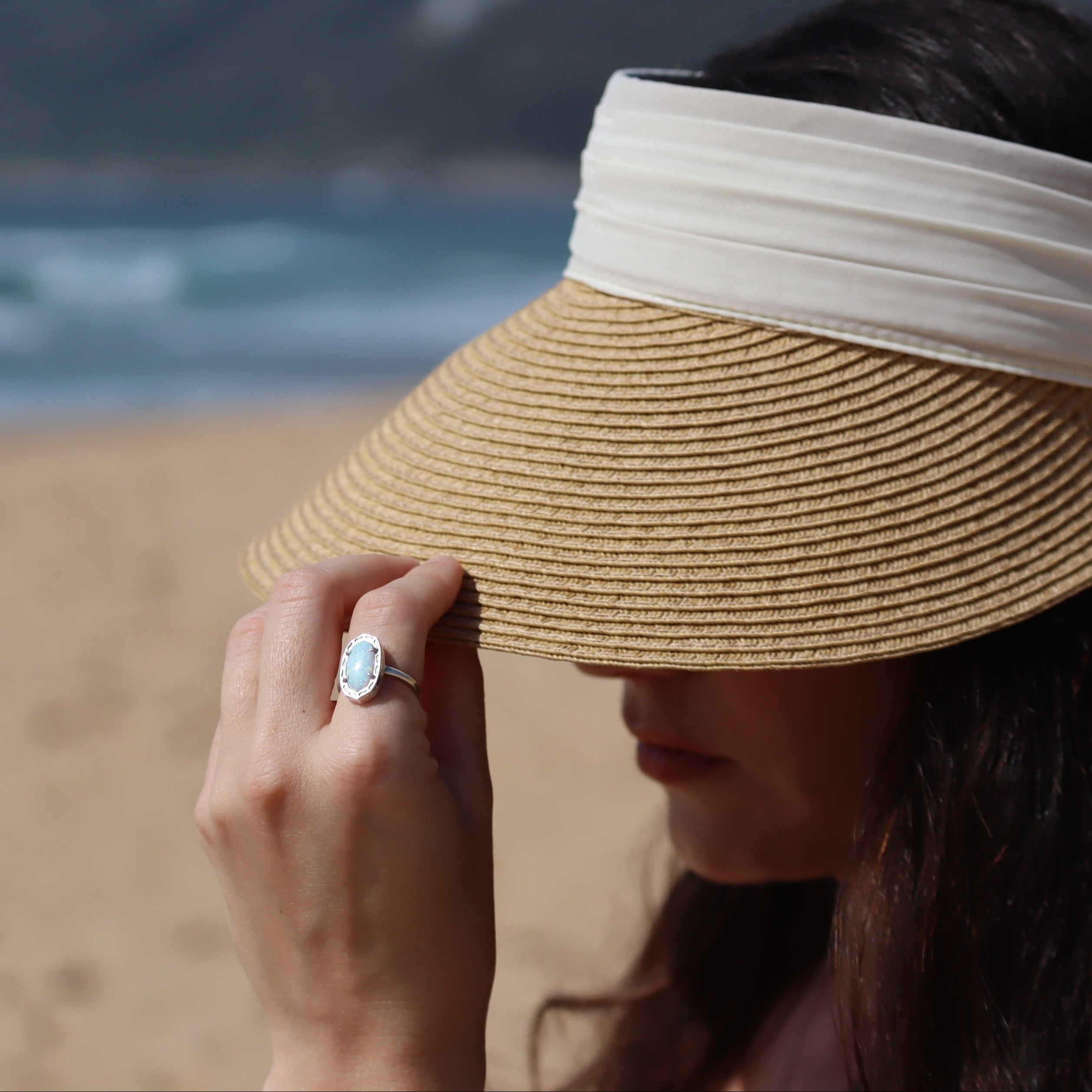 Person wearing a straw visor on a beach with ocean in the background wearing the Olivia Ring set in white lab-created opal