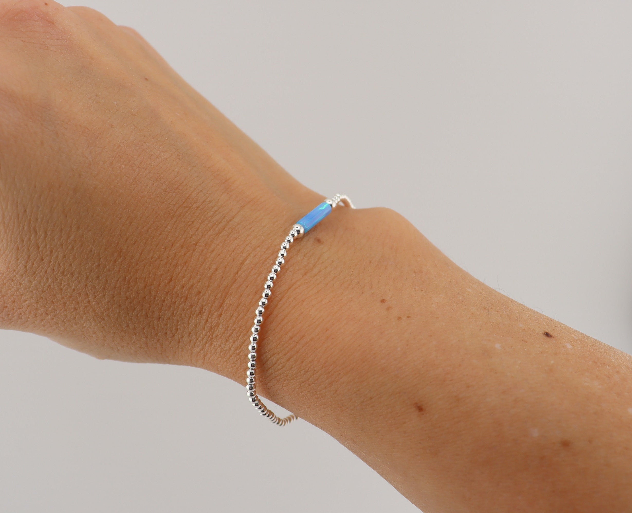 Wrist wearing a silver beaded bracelet with one blue cylindrical bead on a plain white background