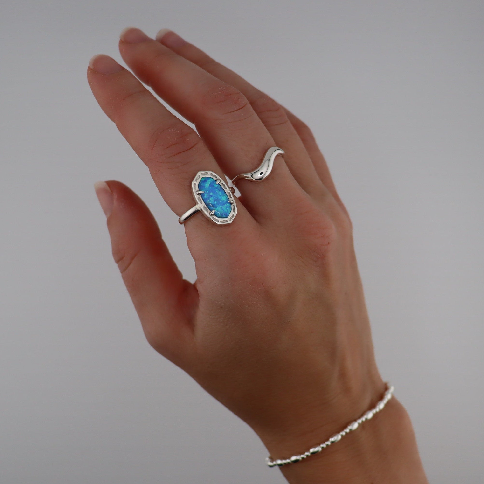 Hand wearing a ring with a blue gemstone and one wavy silver ring on a plain background