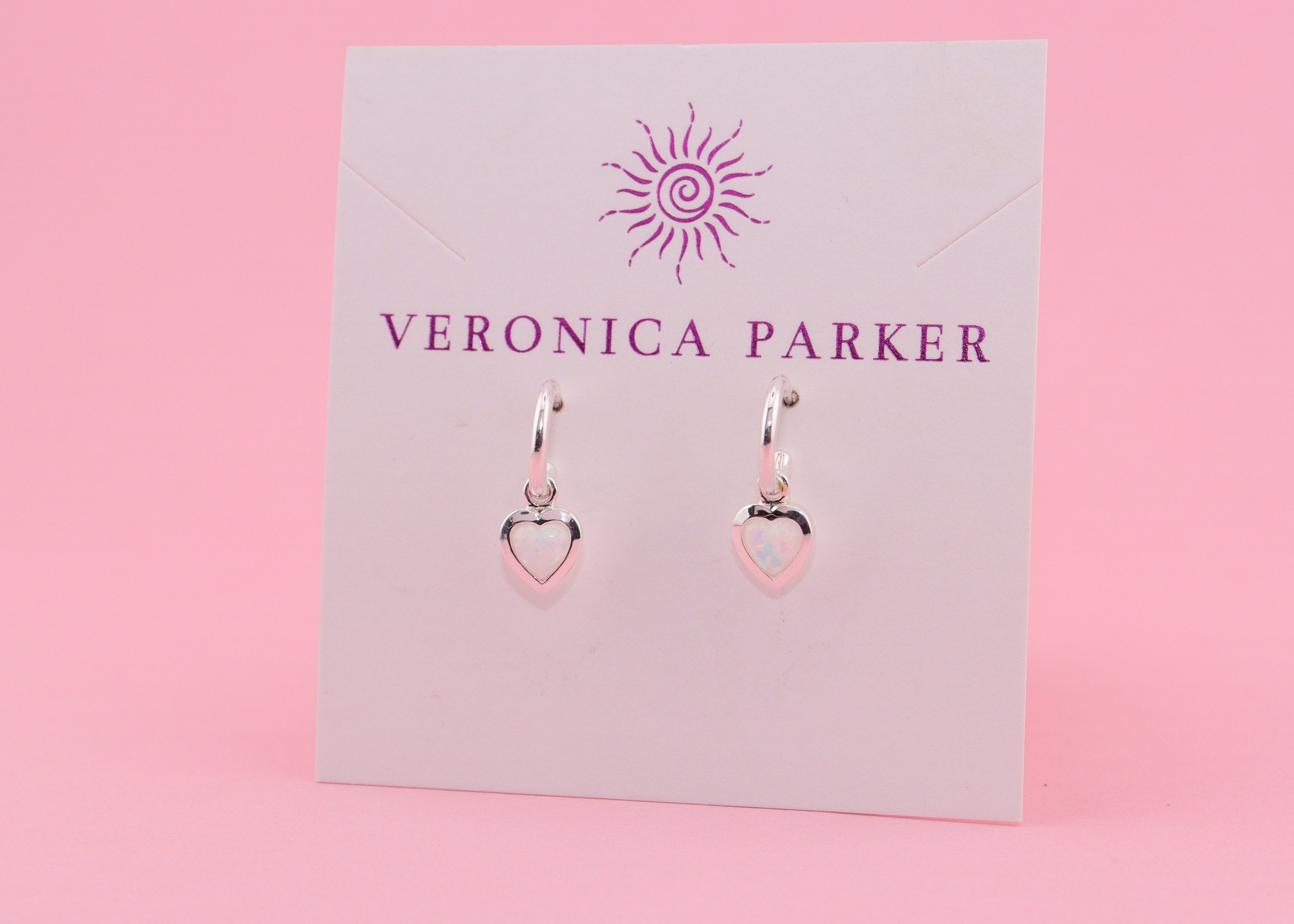 Heart-shaped earrings with white lab-created opals on a Veronica Parker card with a pink background